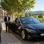 Un chauffeur de VTC ouvre la porte d'une Tesla noire pour un client dans un village provençal ensoleillé, avec vignobles et collines en arrière-plan.